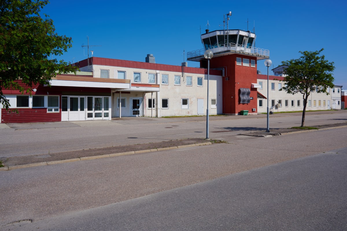 En byggnad med fasader i vitt och rött, och en ljusgrå taklist, syns med en flygledartorn på den högra sidan. Framför byggnaden finns ett asfalterat område med en väg i förgrunden. Två träd är synliga på varsin sida om bilden under en klarblå himmel.