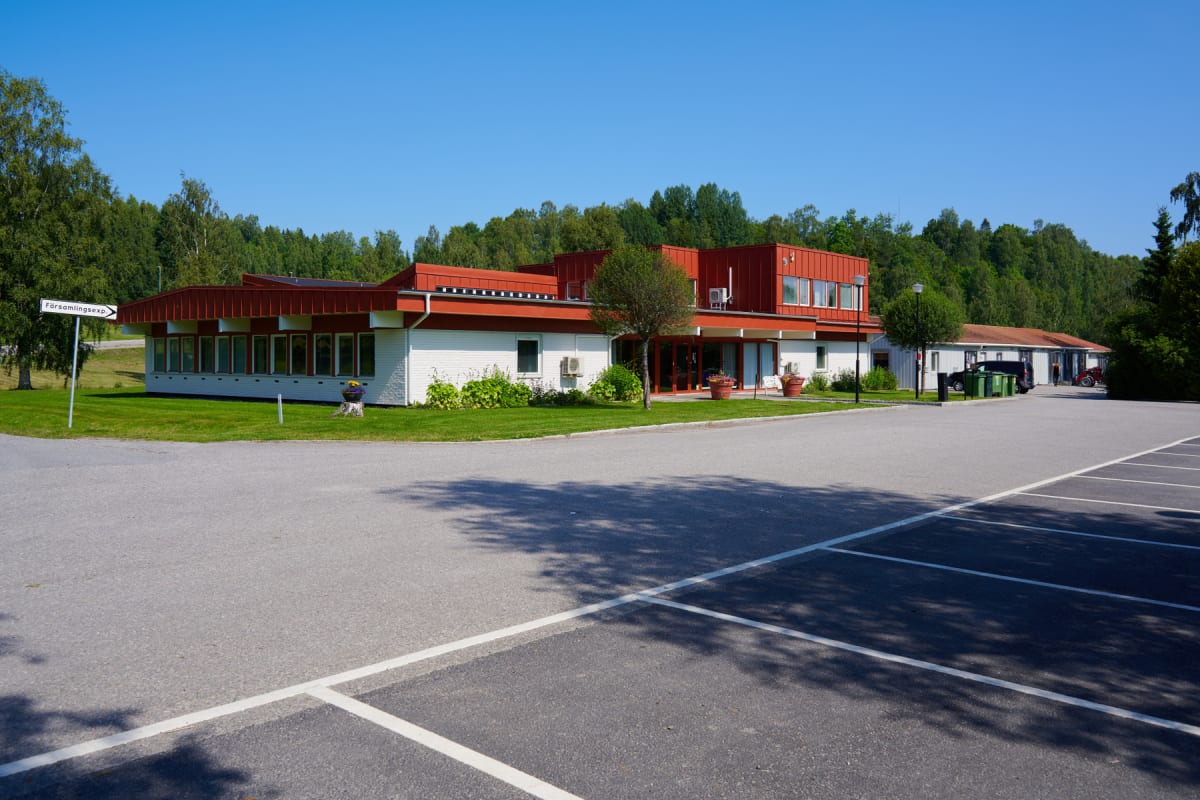 En stor, vit och röd byggnad med en parkeringsplats i förgrunden. Byggnaden har många fönster och en röd taklinje. Runt byggnaden är det gräsmattor och träd. Parkeringsplatsen är to. En skylt med texten "Församlingsexp" syns till vänster.