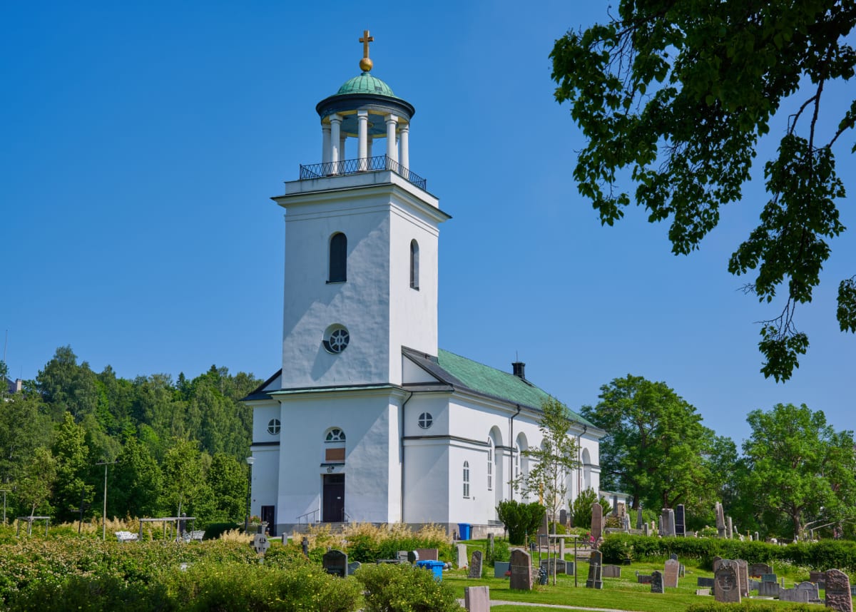 En vit kyrka med grönt tak och en klockstapel med kupol står under en klarblå himmel. Framför kyrkan syns en kyrkogård med gravstenar och grönska, samt träd i bakgrunden. Ett träd med gröna löv ramar in bilden i övre högra hörnet.