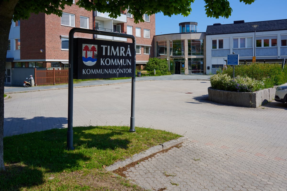 En svart skylt för Timrå Kommun Kommunalkontor står i fokus framför en modern byggnad med glasfasad och en röd tegelbyggnad. En blombänk med vita och lila blommor syns, samt en trottoar. Bladen på ett träd är synliga upptill vänster.