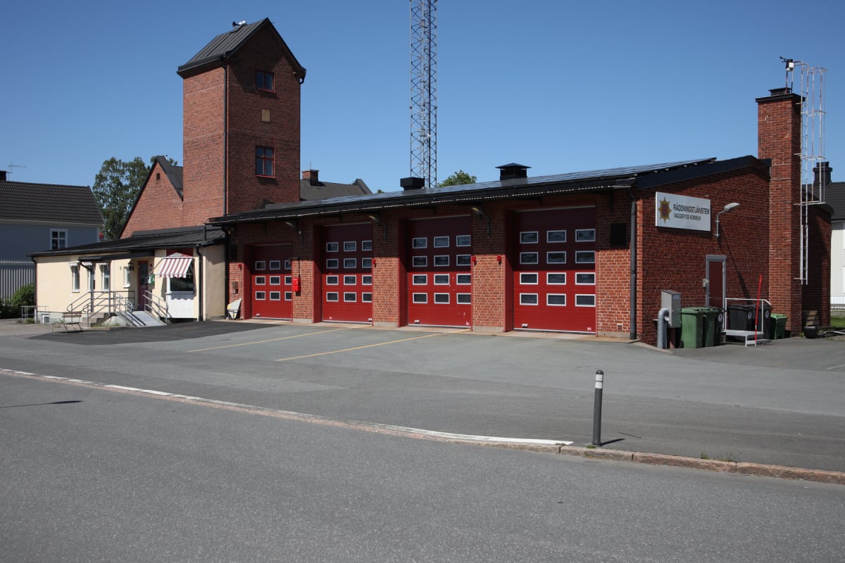 En röd tegelbyggnad med ett högt torn till vänster och flera röda dörrar på framsidan som liknar garageportar. På byggnadens tak finns solpaneler. En skylt med texten 'RÄDDNINGSTJÄNSTEN VAGGeryd KOMMUN' är synlig på väggen. En mast syns i bakgrunden.