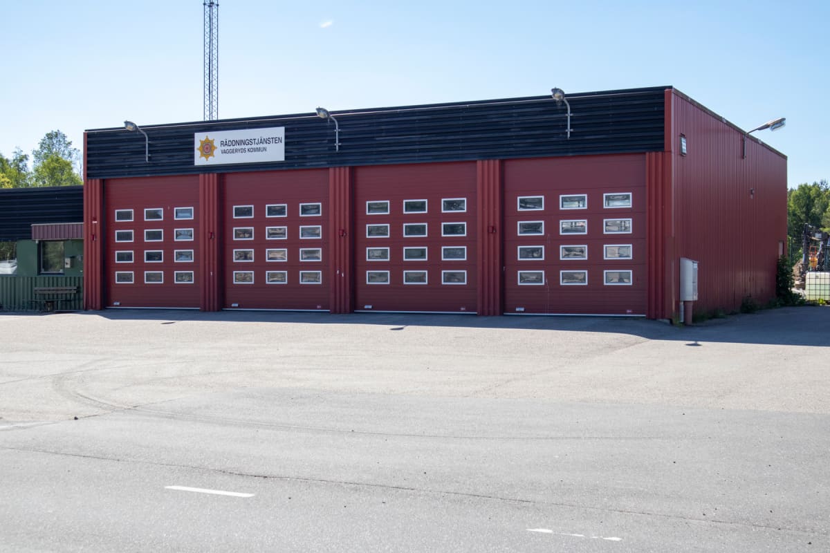 En röd byggnad med fyra stora portar, varav en del av den vänstra syns. Varje port har små fönster uppradade. Ovanför portarna sitter en skylt med texten "RÄDDNINGSTJÄNSTEN VAGGERYDS KOMMUN". Förgrunden består av grå asfalt och i bakgrunden syns skog