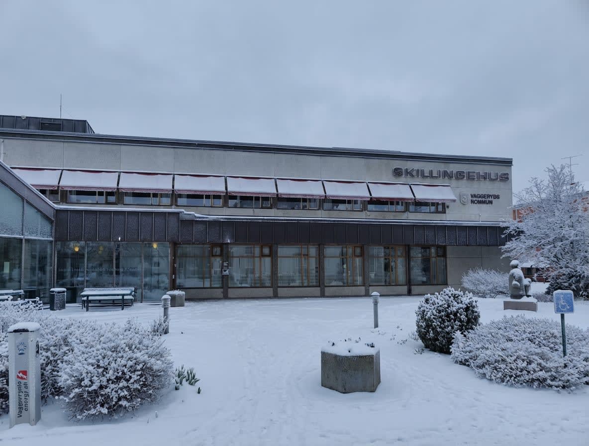 En exteriörbild av en byggnad med snö på marken och buskar. Byggnaden har skylten "SKILLINGEHUS" och "VAGGERYDS KOMMUN". Fönster med röda markiser syns. På gården står en bänk, gatubelysning och en laddstolpe, allt täckt av snö.
