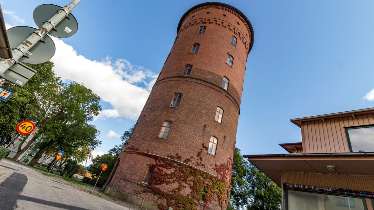 En stor, cylinderformad tegelbyggnad, troligtvis ett vattentorn, sträcker sig upp mot en klarblå himmel med vita moln. Byggnaden är bevuxen med rödaktiga klätterväxter. I förgrunden syns en körbana, vägskyltar och träd.