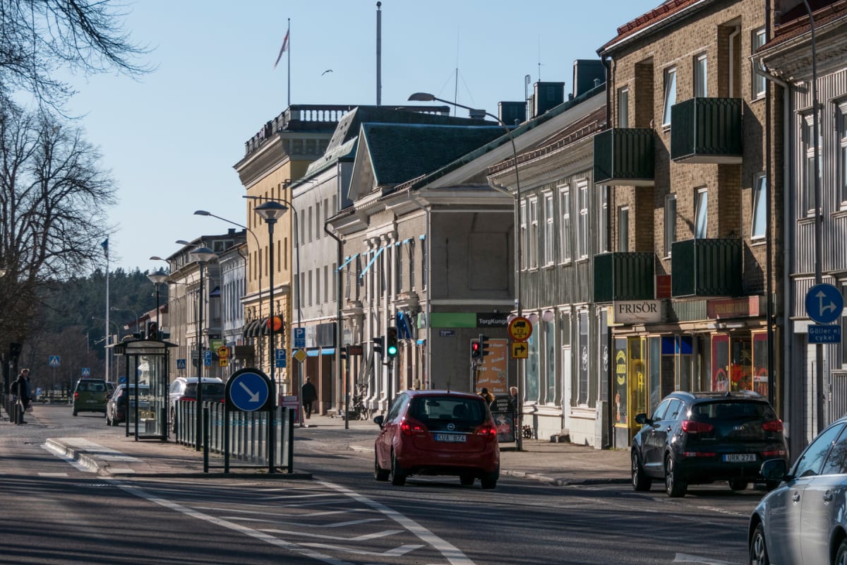 En gata med flera byggnader längs sidorna under en solig dag. Flera bilar är synliga på vägen, varav en röd och en svart bil med synliga registreringsskyltar. Det finns skyltar för olika butiker och en frisörsalong. En flagga syns på taket av en bygg