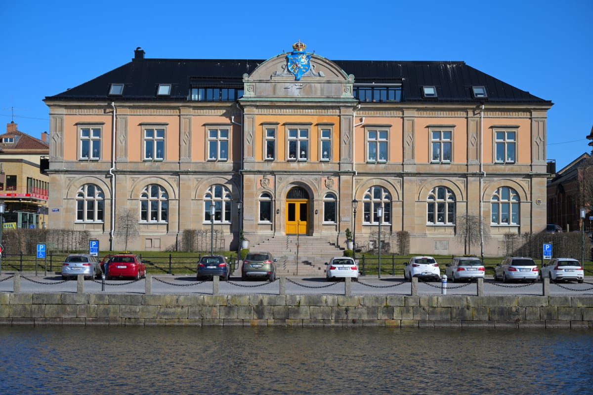 En stor, ljusbrun byggnad med svart tak och gula detaljer i fasaden, inklusive en gul dörr. Framför byggnaden finns en parkeringsplats med flera parkerade bilar och en mur som avgränsar till en vattenyta i förgrunden. Blå himmel ovanför.