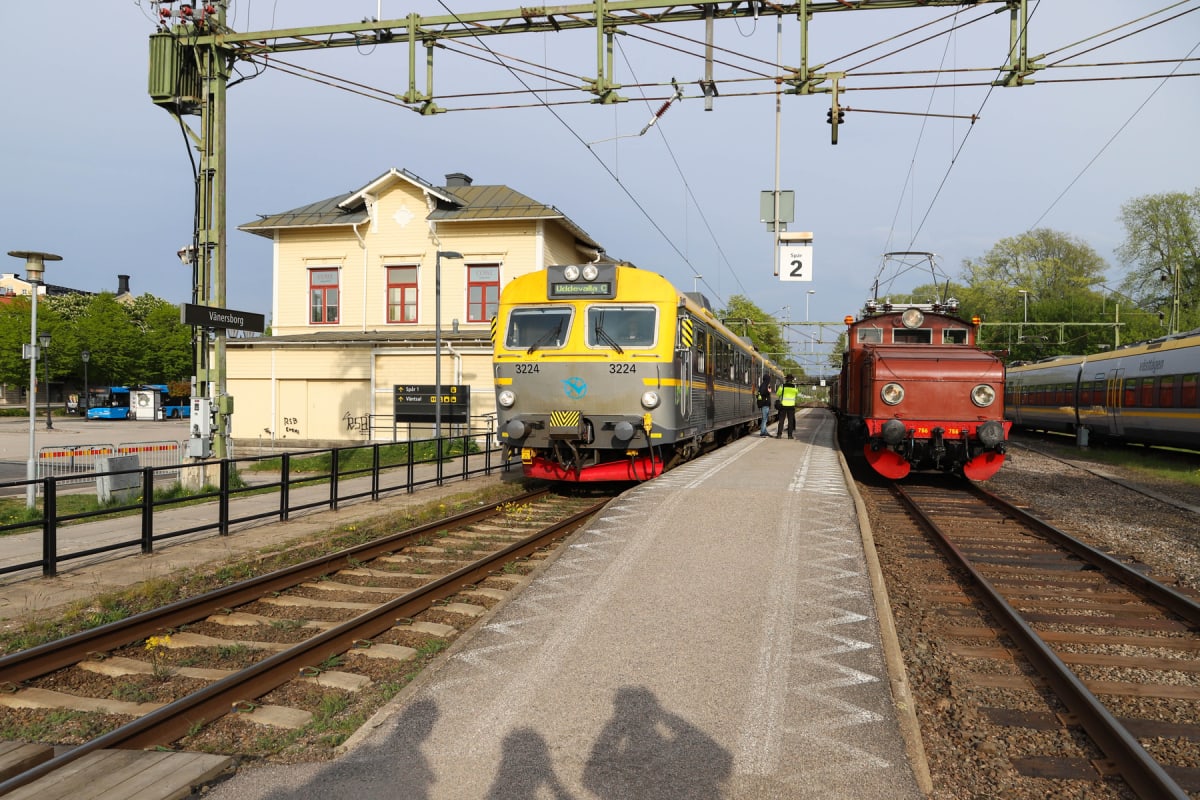 En gul och grå motorvagn med nummer 3224 står på ett spår bredvid en plattform, med en röd lokomotiv 766 på ett intilliggande spår. I bakgrunden syns en gul stationsbyggnad och skyltar som visar "Vänersborg" och "Spår 2". Ovanför spåren finns kontakt