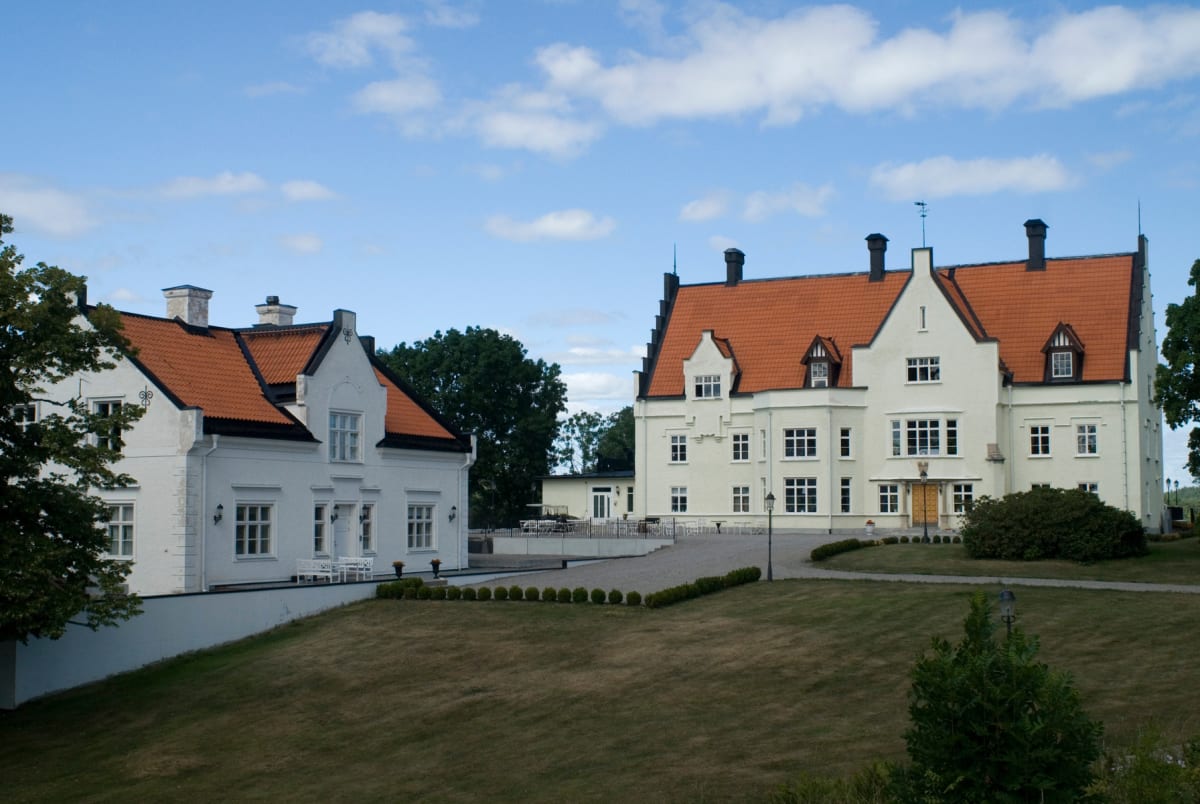 En bild som visar två stora, ljusa byggnader med röda tak under en blå himmel med vita moln, omgivna av grönska och en gräsmatta. Byggnaderna har många fönster och skorstenar, samt ornamenterade detaljer.