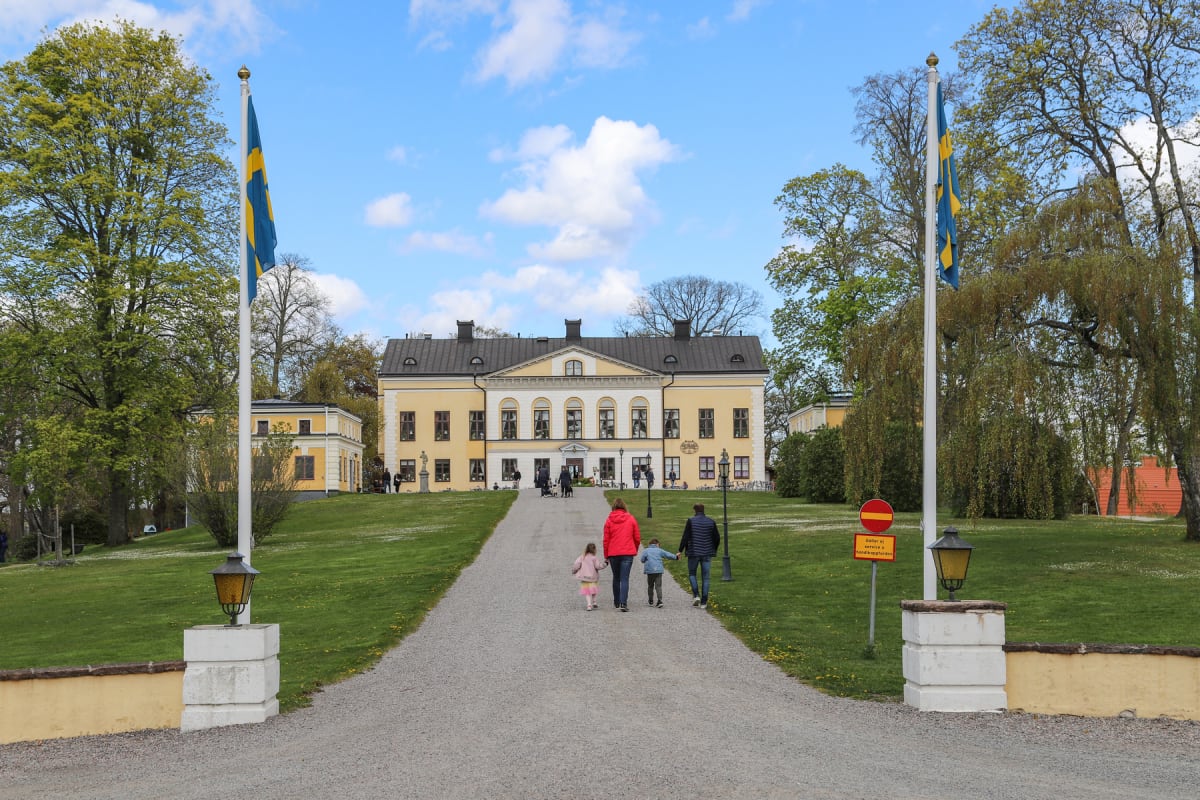 En bild som visar en bred grusgång som leder upp mot en större gul byggnad med vita detaljer och svarta tak. Runt grusgången finns gräsytor och träd med grönska. Två flaggstänger med svenska flaggor står vid början av grusgången. Fyra personer, varav
