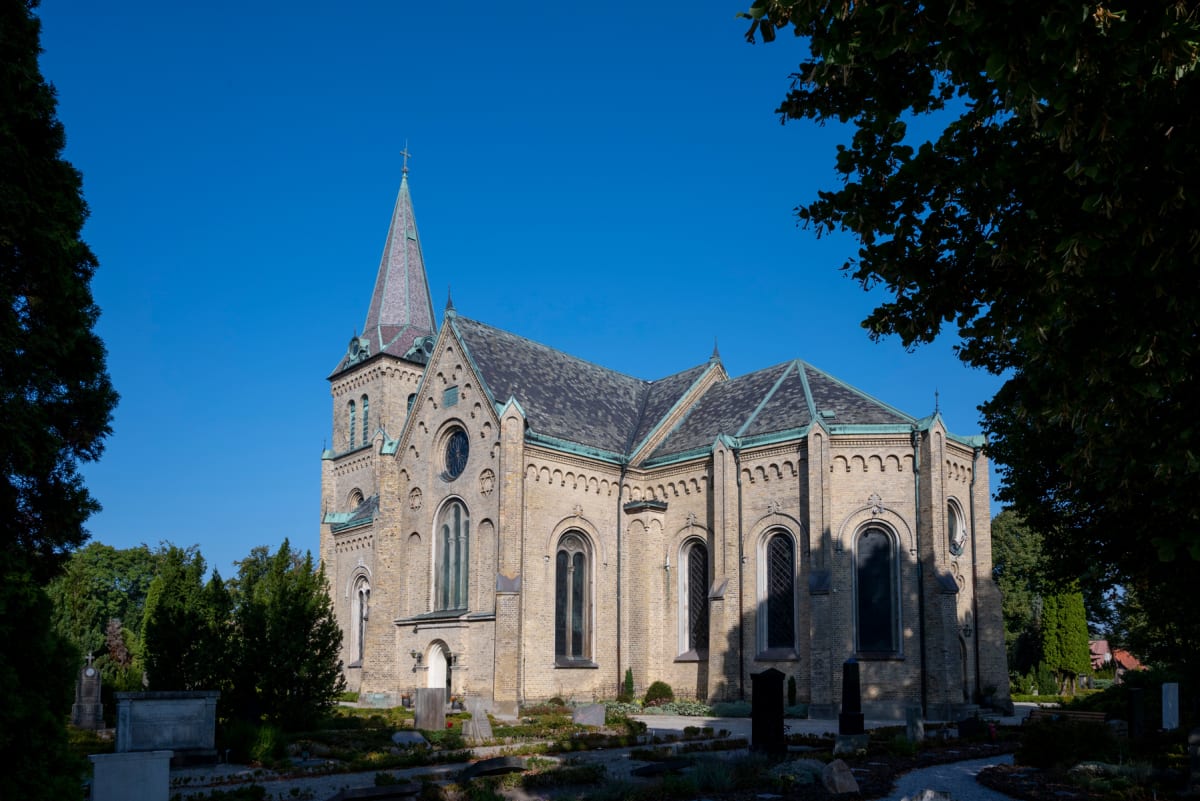 En stor, ljusbrun kyrka med en hög spira står under en klarblå himmel. Runt kyrkan finns en grönskande kyrkogård med gravstenar och träd, varav en del av ett stort träd skymtar i förgrunden till höger.