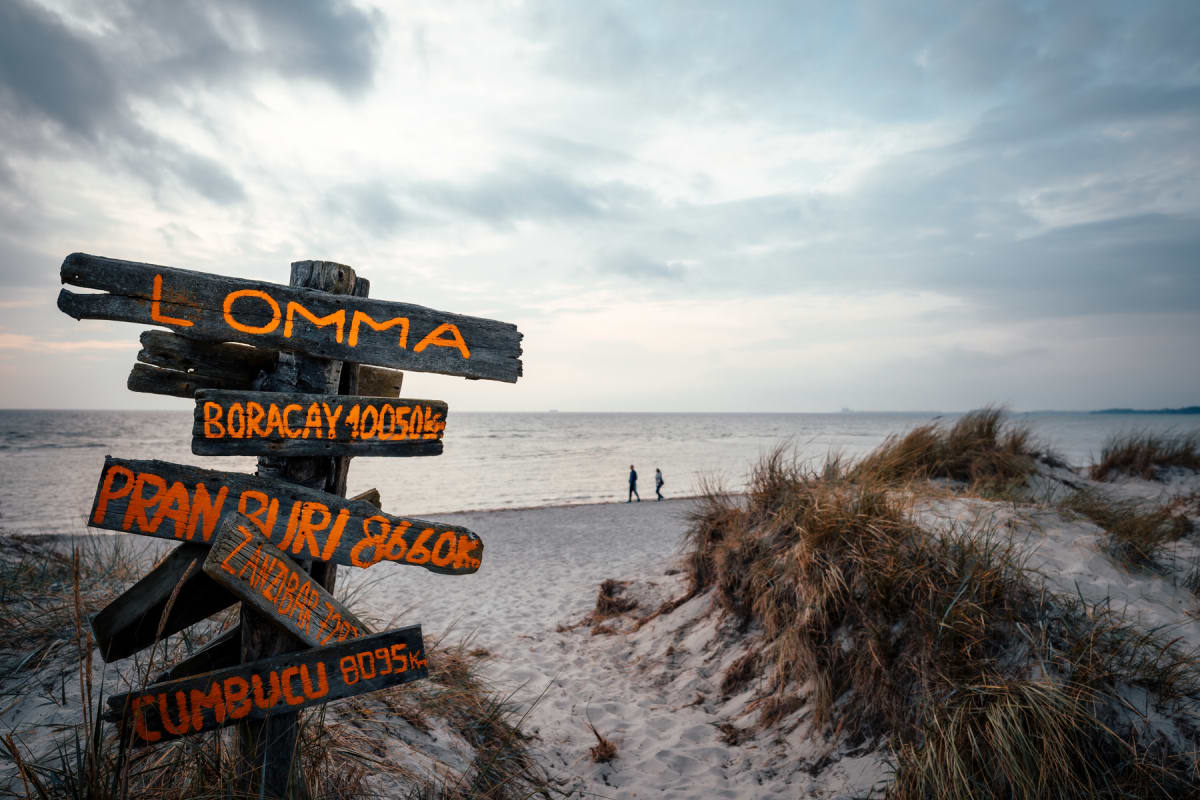 En sandstrand med en handgjord träskylt i förgrunden som pekar ut riktningar och avstånd till olika platser, bland annat Lomma. Två människor syns promenera vid vattenbrynet längre bort. På stranden finns även sanddyner med gräs. Havet och en molnig