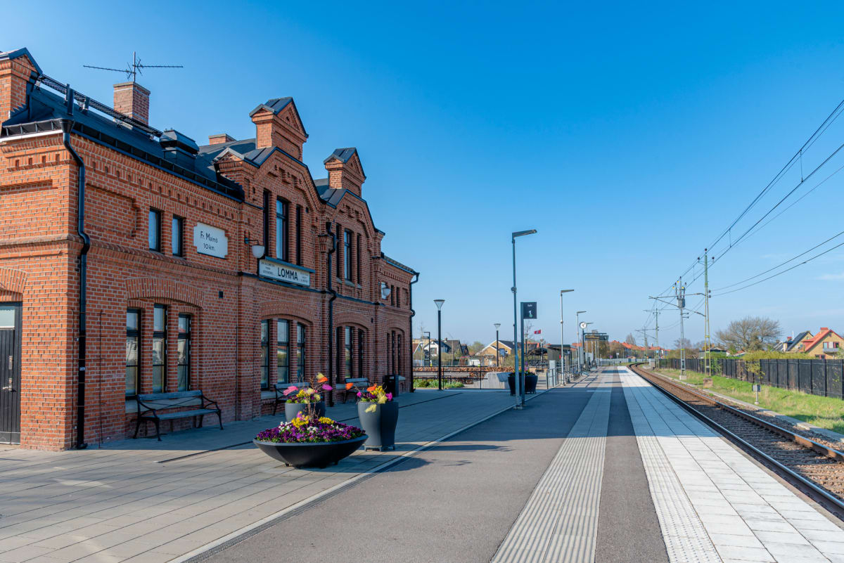 Bilden visar byggnaden för Lomma station en solig dag. Till vänster i bilden syns stationshuset i rött tegel med texten "LOMMA" och stationsnamn. Framför stationshuset finns blomsterurnor och en bänk. Ett spår löper till höger i bild.