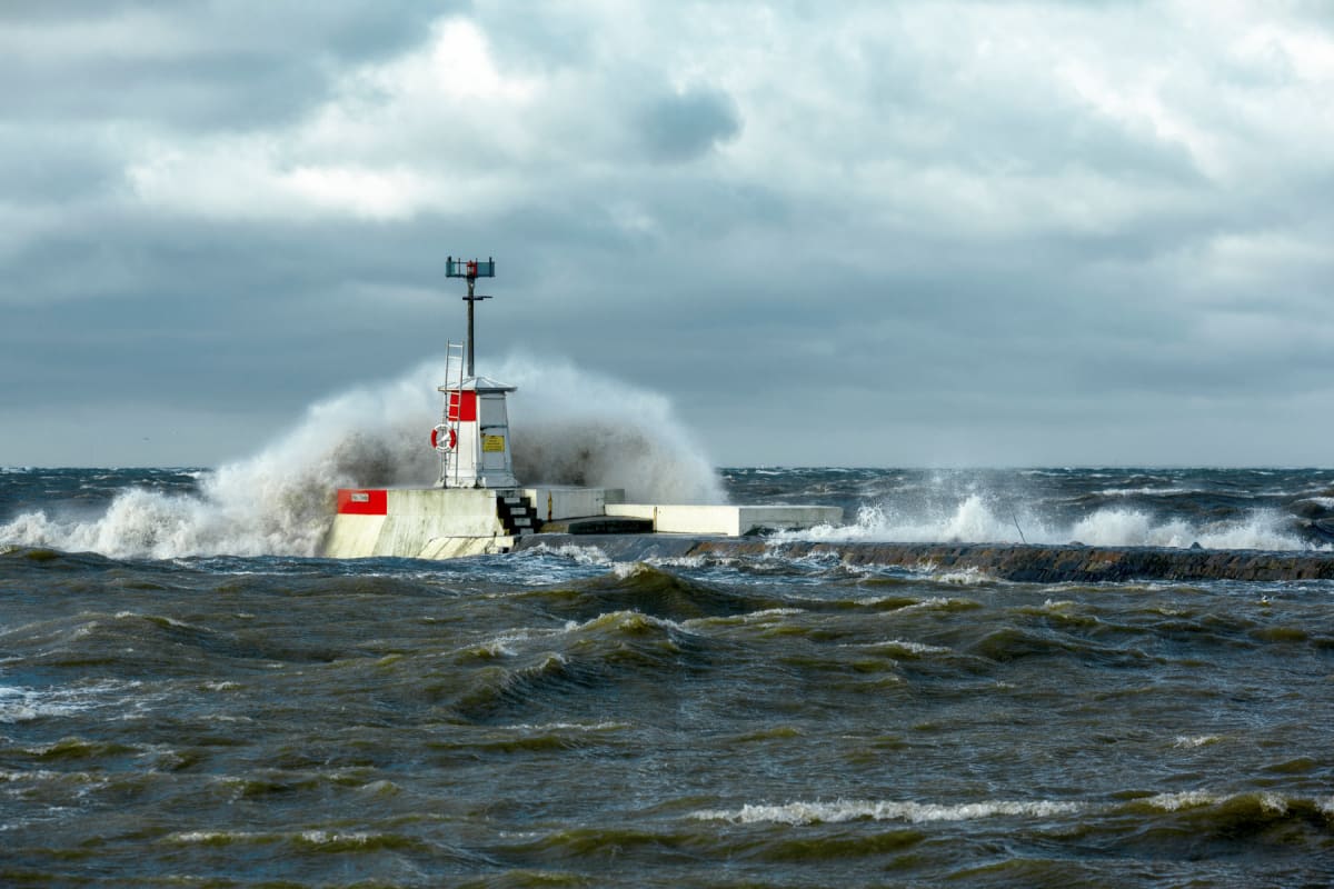 En fyr på en vågbrytare vid ett stormigt hav med vågor som slår mot konstruktionen under en molnig himmel. Fyren är vit med röda detaljer och ett räddningshjul hänger på sidan. Vågorna är mörka och vattnet sprutar upp mot himlen.