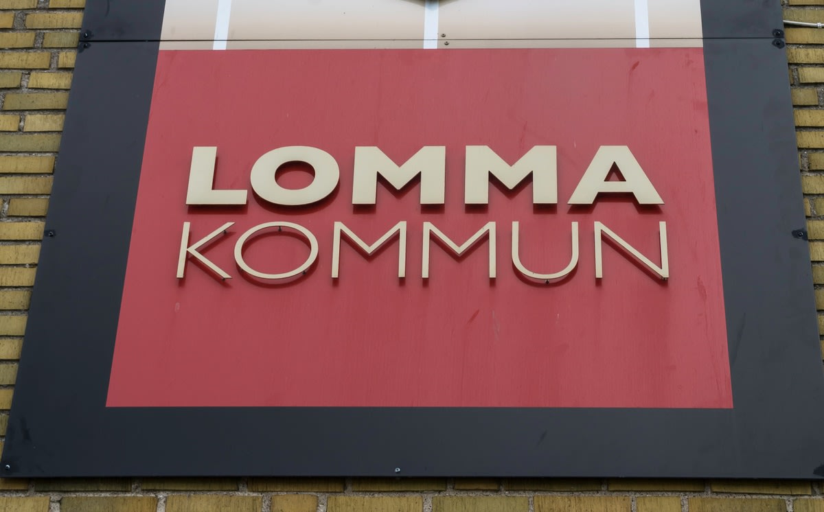 En skylt för "LOMMA KOMMUN" med guldfärgade bokstäver på en röd bakgrund, monterad på en byggnad med gult tegel. Skylten har en svart ram runt den röda ytan. Bilder är tagen snett nedifrån, med tegelväggen i bakgrunden till vänster och nedtill.