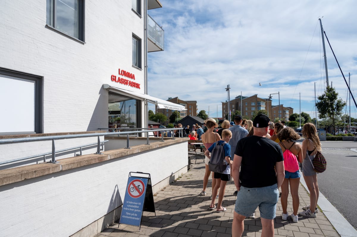 En folksamling framför en byggnad med skylten "Lomma Glassfabrik" under en solig dag. Personerna står i kö på en trottoar invid en väg. I bakgrunden syns segelbåtsmaster, byggnader och träd. En skylt med texten "Vi hänvisar till cykelstället tvärs öv
