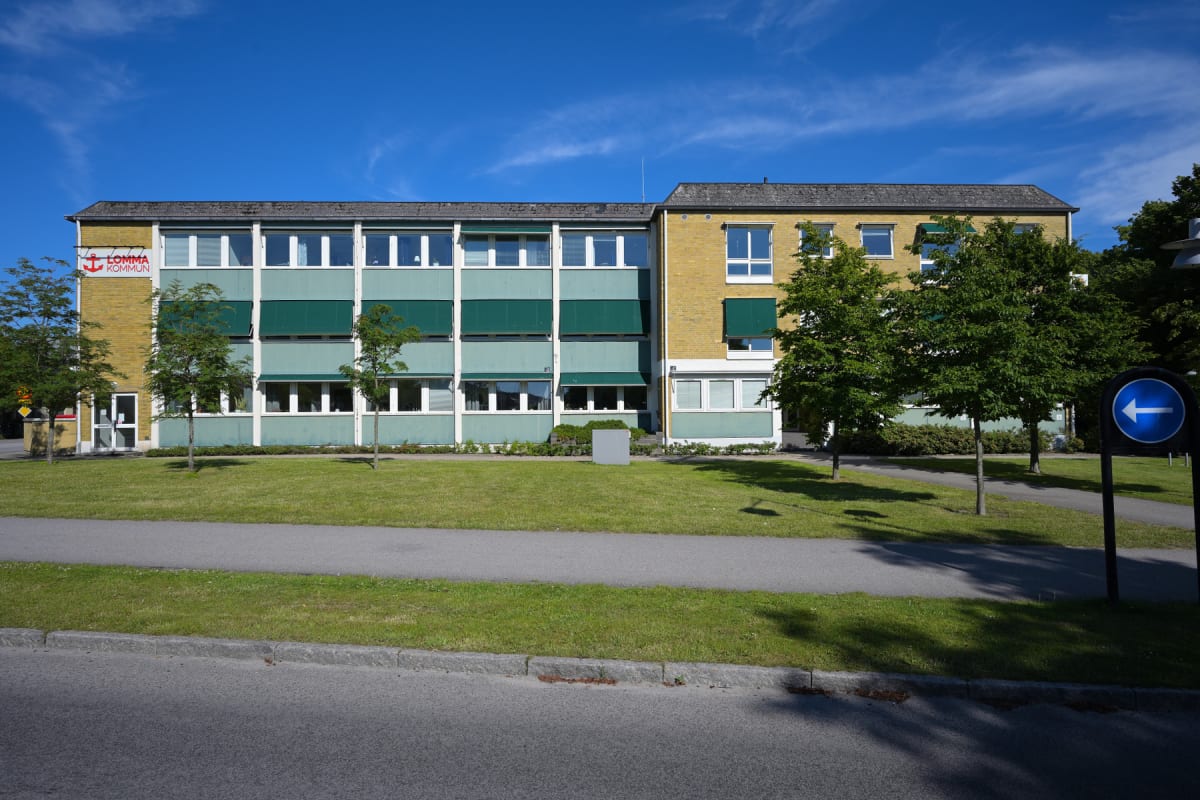 En gul och grön byggnad med flera fönster och mörka markiser, framför vilken det finns en gräsmatta, en gångväg och två små träd. Huset har en skylt med texten 'LOMMA KOMMUN' och en röd ankarelogotyp. En blå vägvisare med en vit pil pekar åt vänster.