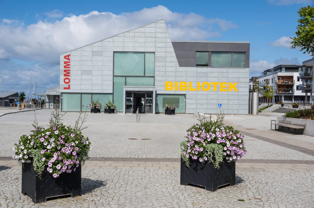 En modern byggnad med bokstäverna "LOMMA" i rött och "BIBLIOTEK" i gult på fasaden, omgiven av kullersten och blomlådor, under en himmel med vita moln. I bakgrunden syns segelbåtsmaster och fler byggnader.