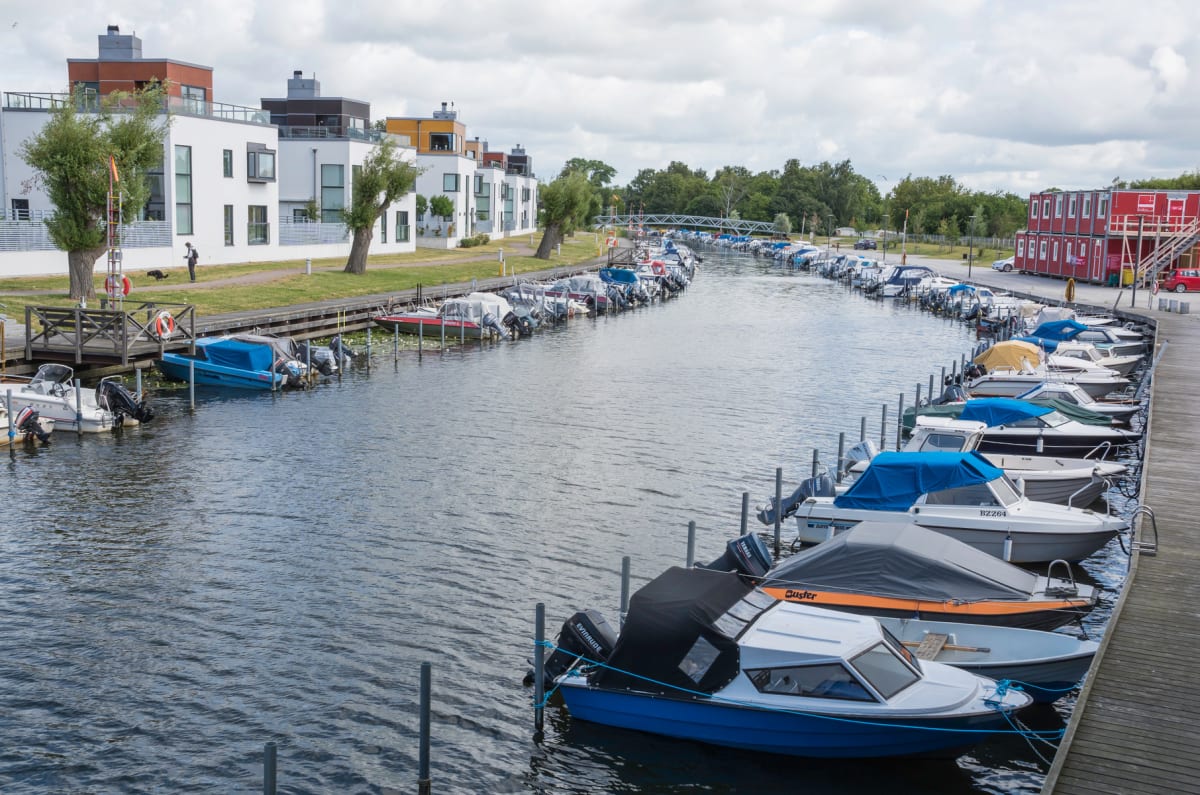 En översiktsbild av en kanal kantad med förtöjda motorbåtar längs båda sidor. På vänster sida syns vita flerfamiljshus med moderna fönster och en gräsplätt vid vattnet. På höger sida finns en träbrygga och bakom den containerliknande röda byggnader.