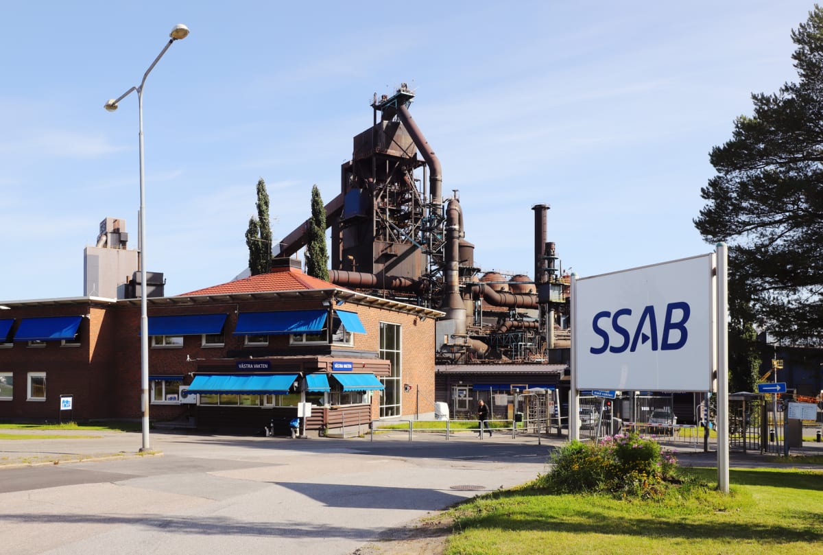 En stor översiktsbild av ett industriområde med en stor SSAB-skylt i förgrunden till höger. Ett stort industribyggnadskomplex med rör och skorstenar dominerar bakgrunden. Ett tegelhus med blå markiser och skylten ”Västra Vakten” syns till vänster.