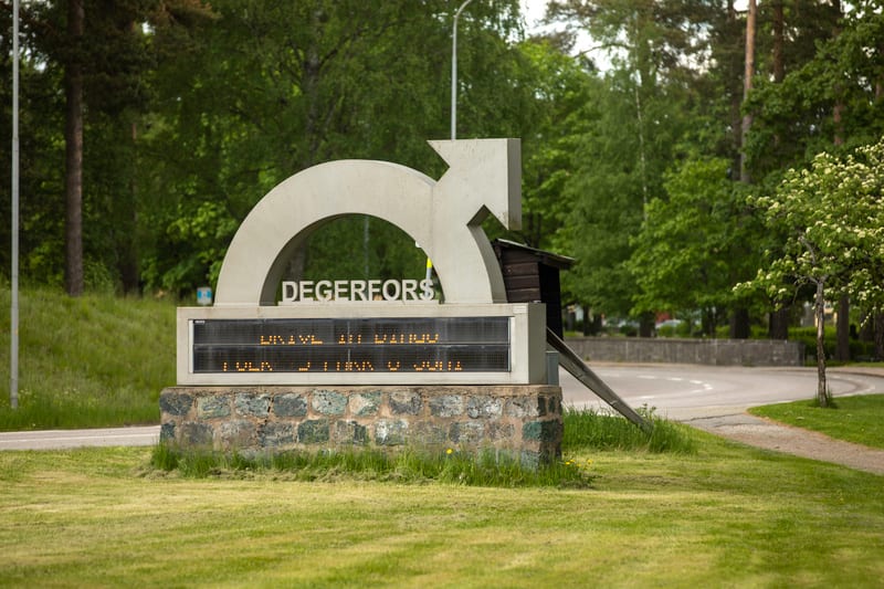 Bilden visar en skylt med texten "DEGERFORS" placerad på en stenbas. Under texten finns en digital display med gulaktig text som till stor del är oläslig men visar delar av ord som "UNIVE" och "JULI". Skylten har en bågformad del och en pilformad del