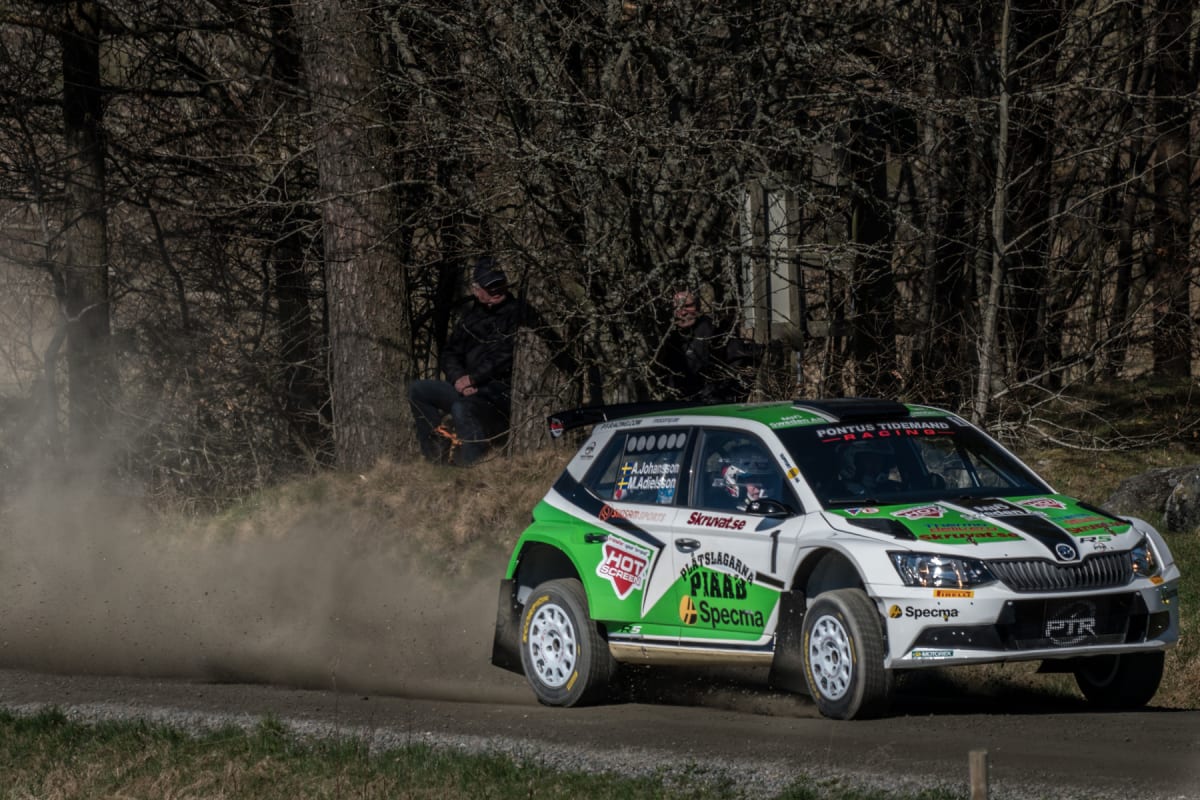 En vit och grön rallybil kör på en grusväg och skapar ett dammoln. I bakgrunden syns träd utan löv och två personer sittande vid skogsbrynet. Solljuset faller in från höger.