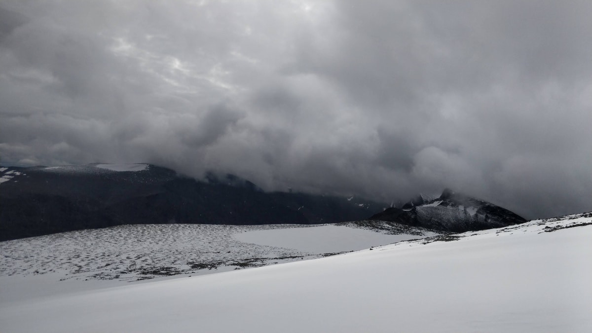 En vidvinkelbild tagen utomhus visar ett bergslandskap vid Kebnekaise med snötäckta ytor i förgrunden och ett mörkt bergsparti i bakgrunden. En disig, molnig himmel täcker en stor del av bergstopparna.