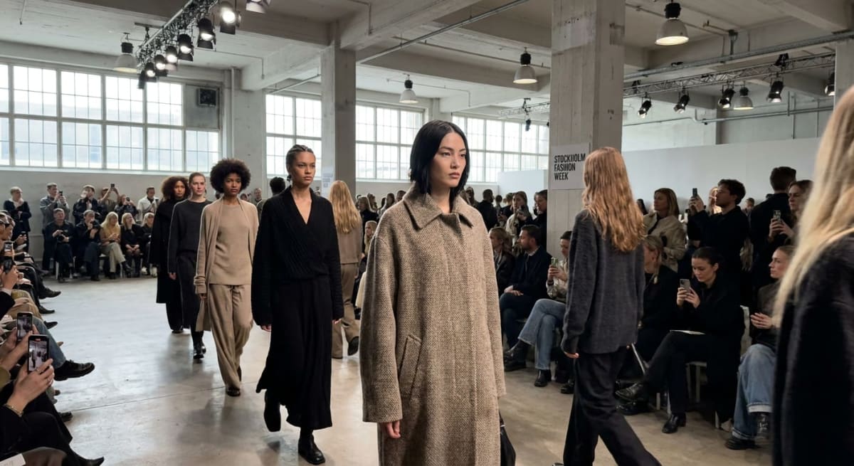 En grupp modeller går längs en bana under en modevisning. Till vänster i bilden syns åskådare som fotograferar evenemanget med mobiltelefoner. En skylt med texten "STOCKHOLM FASHION WEEK" syns i bakgrunden. Takbelysningen syns tydligt.