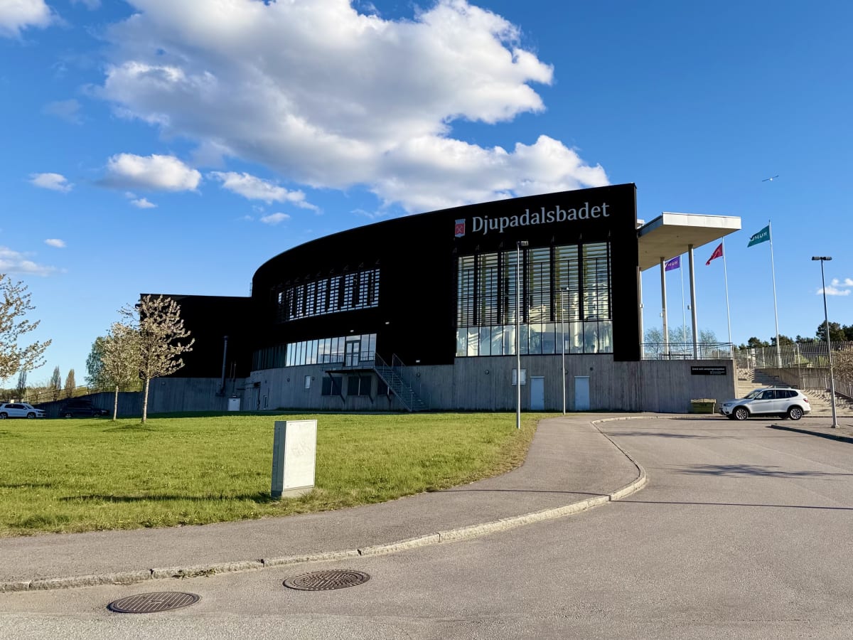 En modern svart byggnad med stora fönster och texten "Djupadalsbadet" ovanför huvudentrén. Flaggstänger med flaggor i rött, lila och grönt syns bredvid byggnaden. Framför byggnaden ligger en grön gräsmatta och en asfalterad gångväg under en blå himme