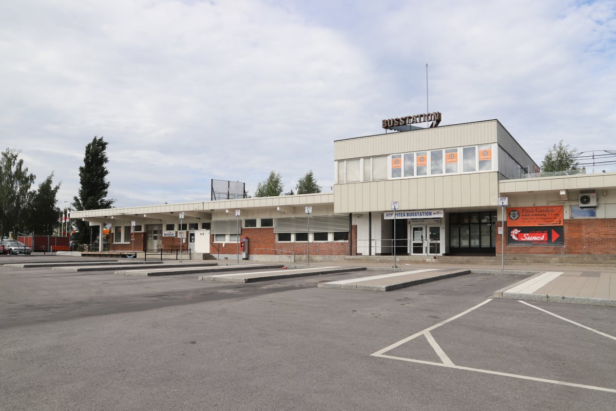 En bild som visar en bussstation under dagtid. Byggnaden är en modern tegelbyggnad med ett "BUSSTATION"-skylt på taket. Framsidan har flera fönster och en entré. En asfalterad bussplan med upphöjda markeringar för bussar ligger framför stationen. Sky