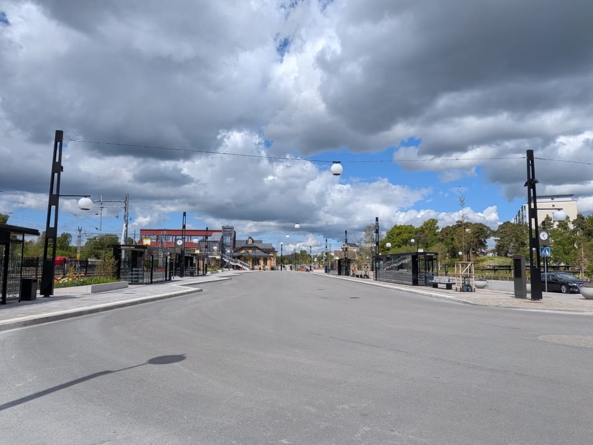 En gata framför en byggnad under en delvis molnig himmel. Längs gatan kantar ljusstolpar och busshållplatser. På håll syns en byggnad med en trappa och ytterligare byggnader i bakgrunden. En svart bil står parkerad till höger i bilden.