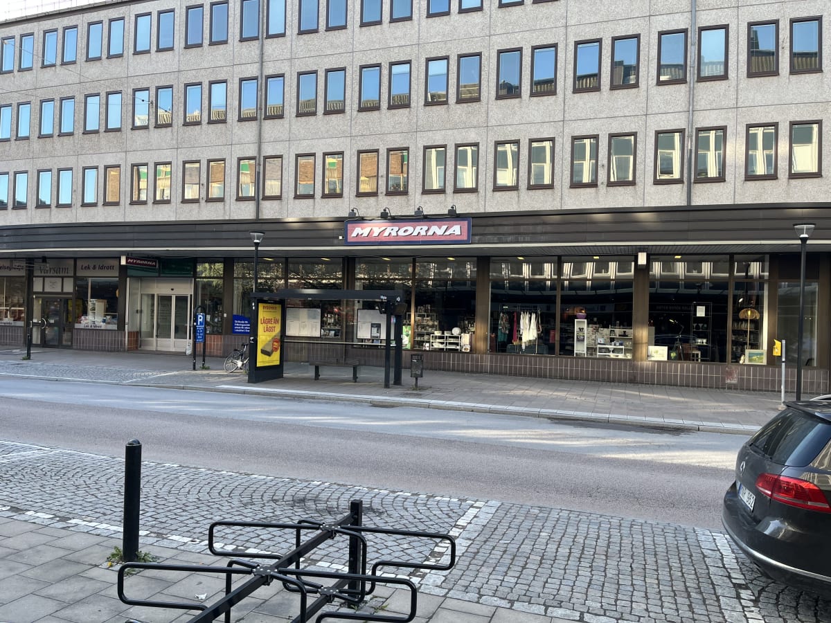 En gata med gatsten och asfalt i förgrunden, parkeringsplatser, en busskur, och kommersiella byggnader i bakgrunden. Butiker som "Värsam", "Lek & Idrott" samt "Myrorna" är synliga. Ett svart fordon skymtar i högra kanten.