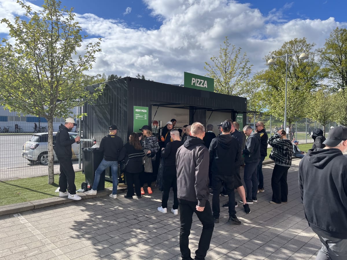 En utomhusbild föreställande en kö med minst 15 personer framför en svart matservering med skylten "PIZZA" och "MENY PIZZA" med priser för pizzaslice, Coppersmith 3.5% och läsk. Personerna står på en stensatt yta med grön gräsmatta intill och träd me
