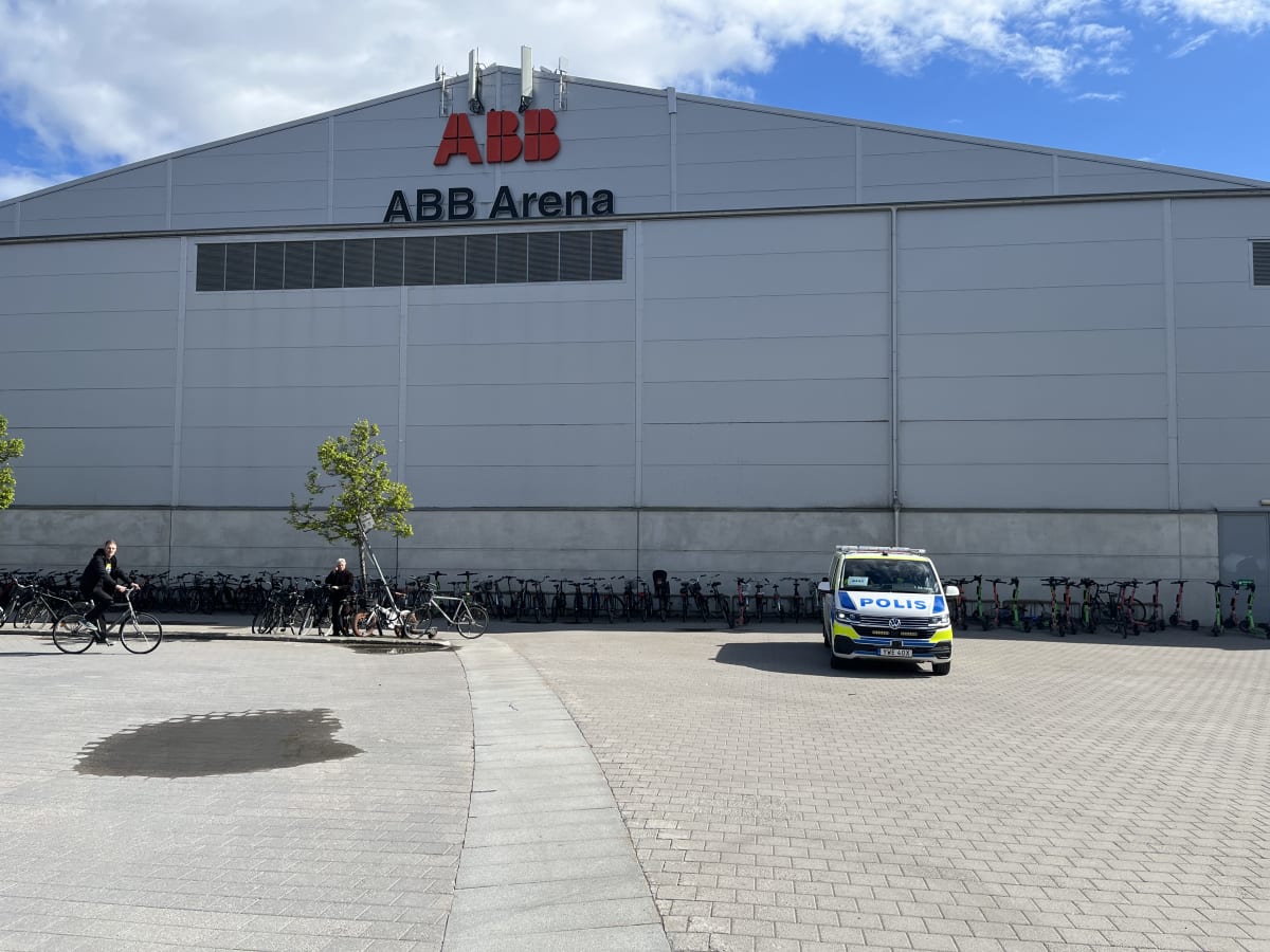 En stor, grå byggnad med texten "ABB ABB Arena" på fasaden dominerar bilden. Framför byggnaden står en svensk polisbil parkerad. Till vänster i bilden cyklar en person förbi och i bakgrunden syns många uppställda cyklar och elsparkcyklar. Molniga him