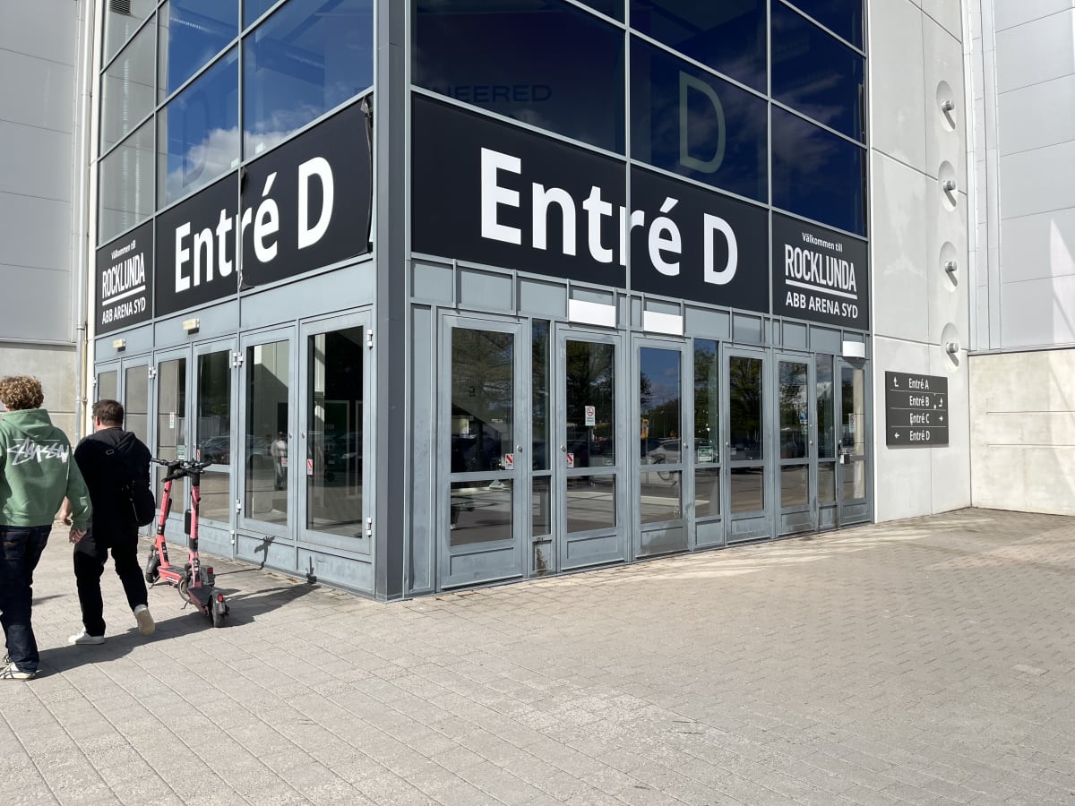 En bild som visar byggnaden Rocklunda ABB Arena Syds Entré D. Två män går mot entrén, varav en har en rosa elsparkcykel vid sin sida. Byggnaden har stora glaspartier och svarta skyltar med vit text. I bakgrunden syns en blå himmel med moln.