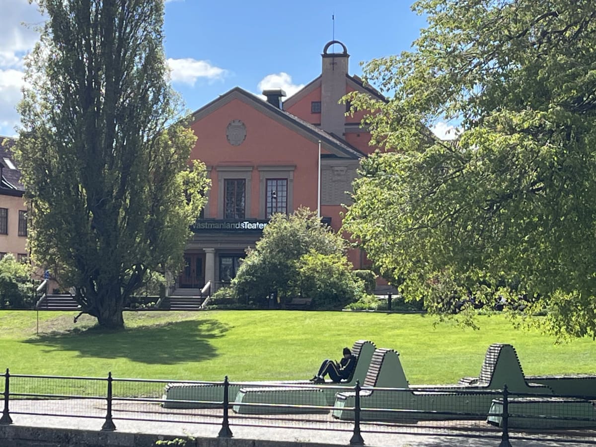 En bild tagen utomhus under dagtid som visar en röd byggnad med texten "Västmanlands Teater" omgiven av träd och en gräsmatta. I förgrunden sitter en person på en grön bänk invid ett staket intill vad som ser ut att vara en kaj eller mark som gränsar