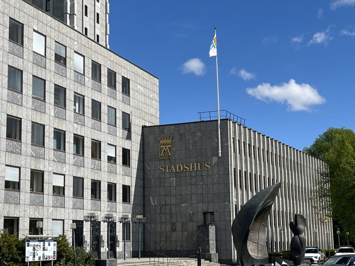 En stor, modern byggnad i grå sten med många fönster. Den har texten "STADSHUS" i guld bokstäver och en flagga på taket. På framsidan syns en skulptur och lövträd, samt en klarblå himmel med några moln.