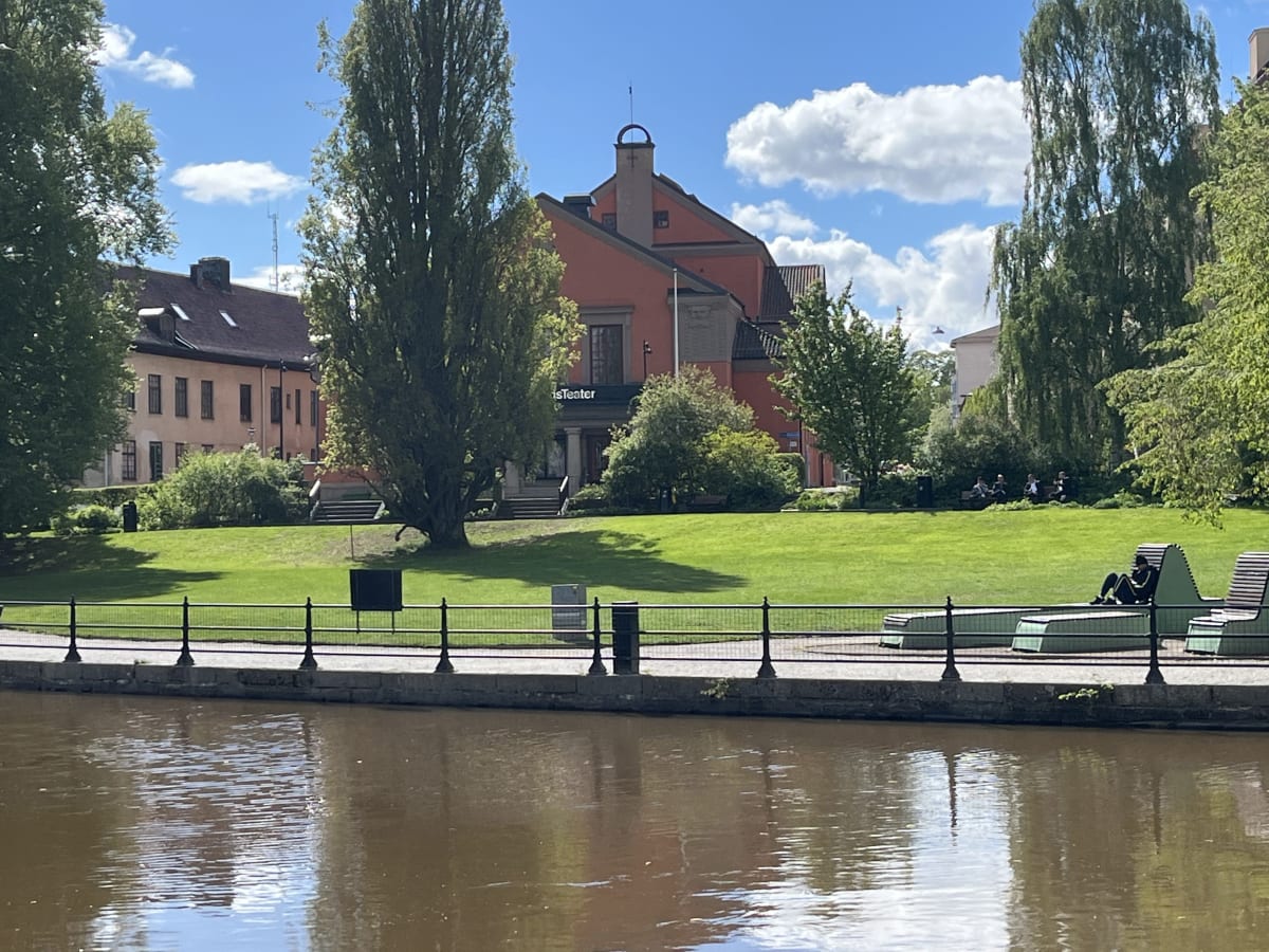 En bild som visar en flod eller å med en stadsmiljö intill. På höger sida syns en park med gräsmatta, träd och bänkar. Till vänster i bilden syns en byggnad med en inskription som lyder "isTeater". Soligt väder med blå himmel och vita moln.