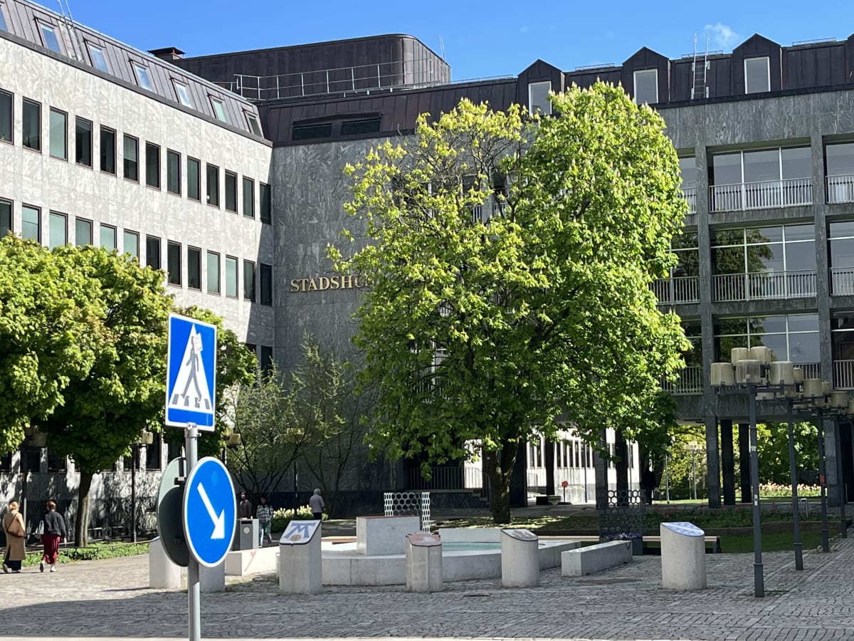 En utomhusbild som visar en stor byggnad med texten "STADSHUS" på fasaden. Framför byggnaden finns flera träd med gröna löv och två trafikskyltar, den ena föreställer ett övergångsställe och den andra en pil som pekar nedåt åt höger. Grå och ljusa by