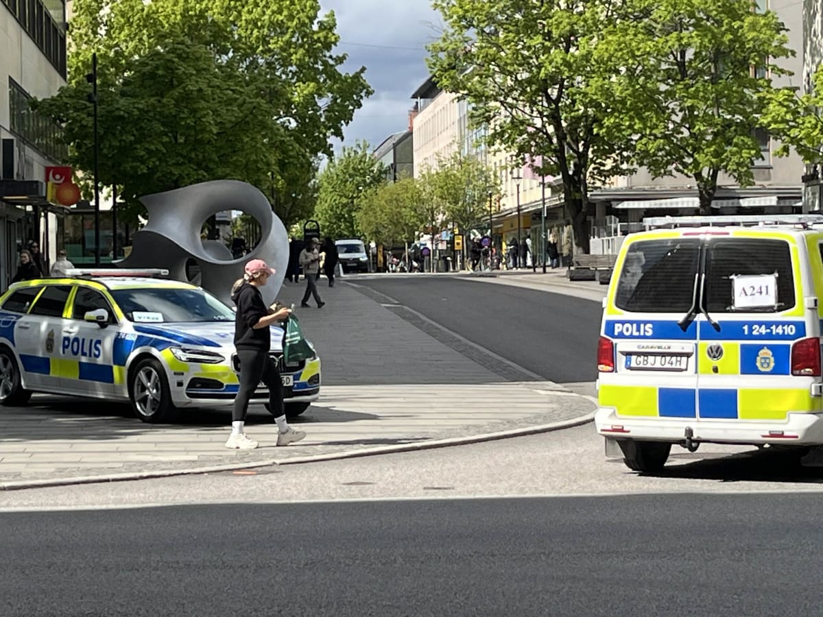 En gata i ett tätbebyggt område med flera personer och två parkerade polisbilar. Till vänster står en Volvo i polismålning och en person går i förgrunden med en påse. Till höger syns bakdelen av en VW skåpbil i polismålning. Gröna träd och byggnader
