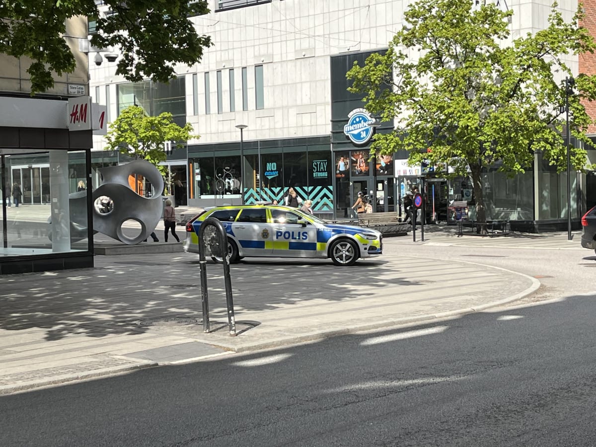 En svensk polisbil står parkerad på en gata i en stadsmiljö under dagtid. I bakgrunden syns butiker, människor, träd med gröna löv och en abstrakt skulptur på ett torg. Solen skiner och skapar skuggor på marken.