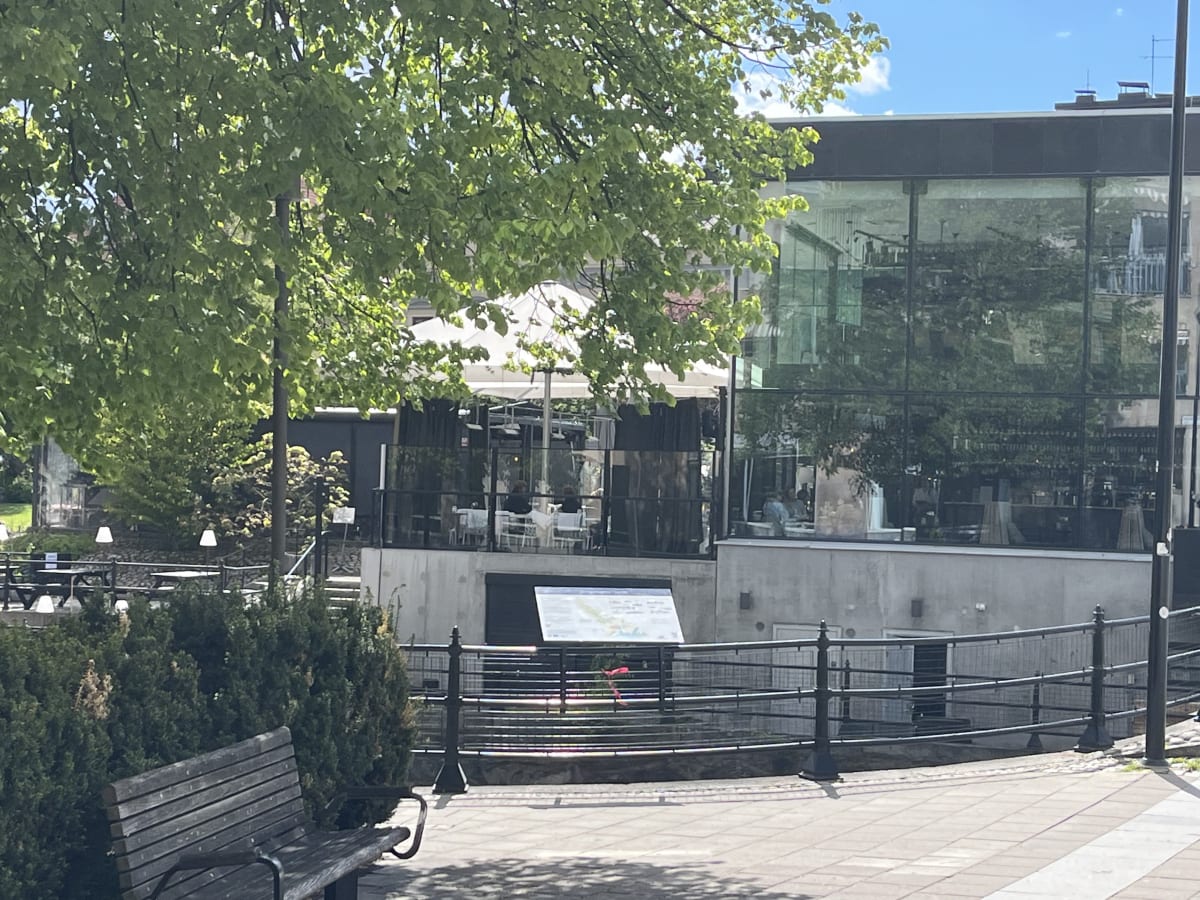 En utomhusbild som visar en modern glasbyggnad med en uteservering. Gröna träd och buskar skymmer delvis byggnaden. En träbänk och ett staket syns i förgrunden, samt ett stenbelagt område. En informationsskylt står nära staketet.