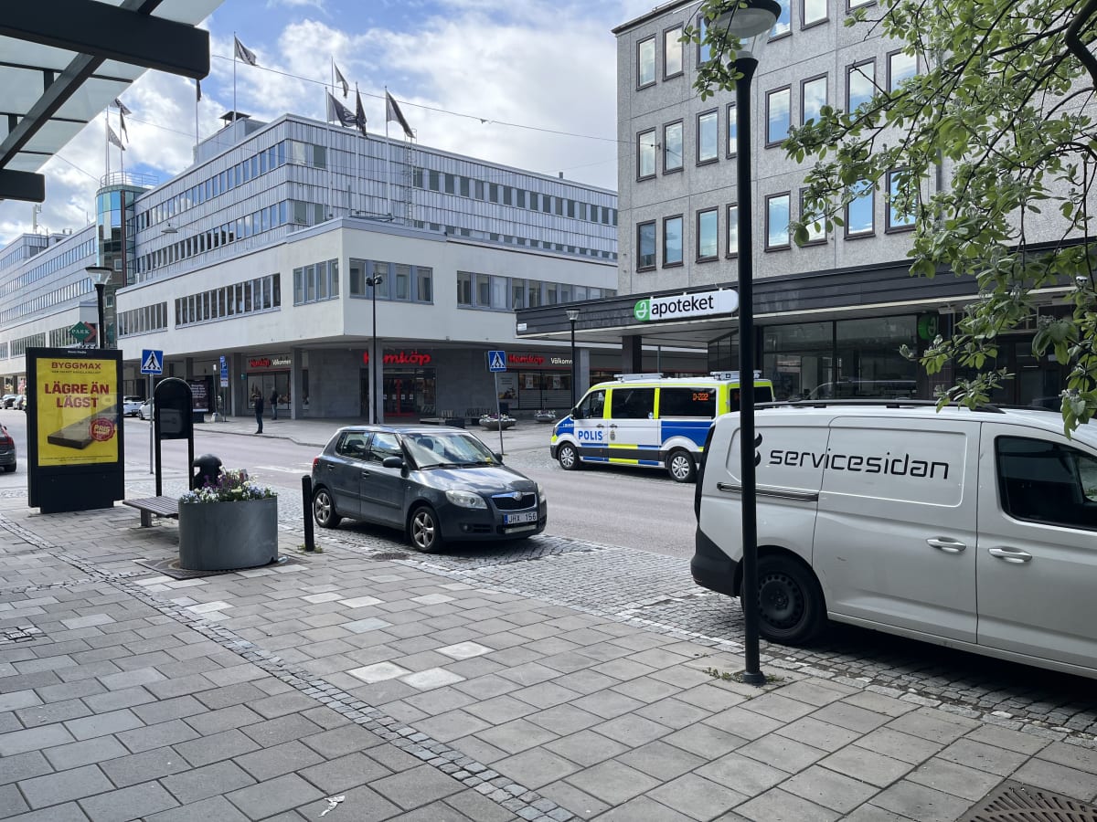 En gatuscen med byggnader, fordon och människor. En polisbil, en grå bil, och en vit skåpbil är parkerade eller står stilla i bilden. På höger sida syns en busshållplats och en bred trottoar med gatustenar och en bänk. I bakgrunden syns Hemköp och et