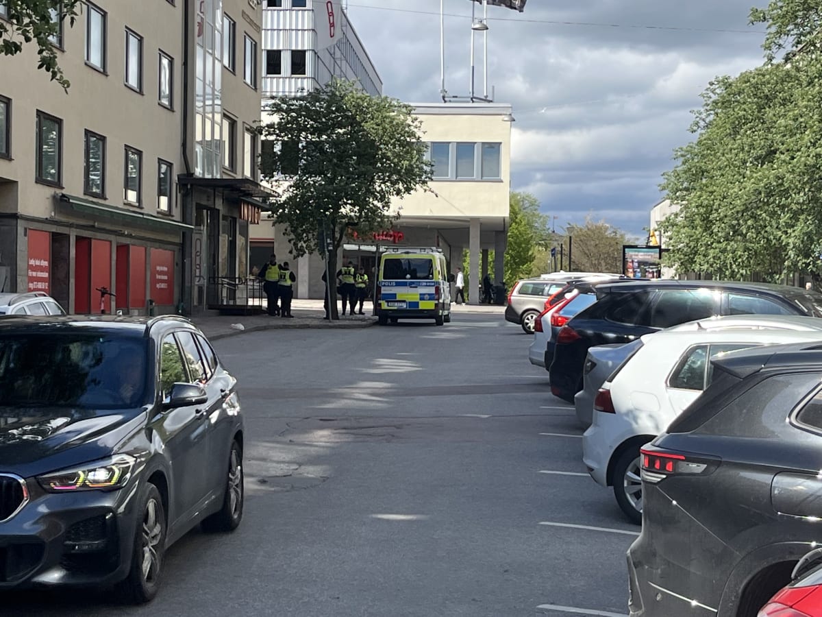En gata med parkerade bilar på höger sida och en byggnad med butiker på vänster sida. I bakgrunden syns en vit polisbil och flera personer, varav några bär gula västar, vid ingången till en byggnad.
