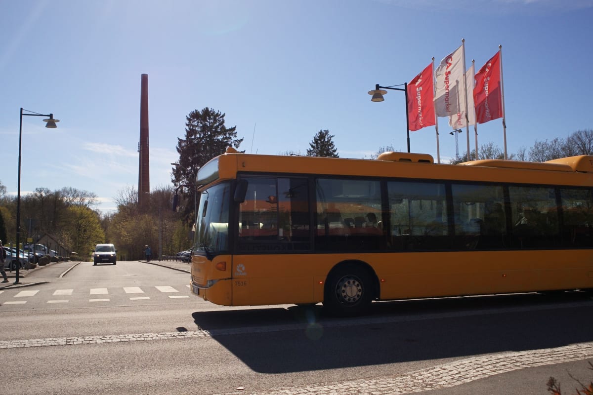 En gul buss kör på en väg med ett övergångsställe en solig dag. I bakgrunden syns skog, en hög tegelpipa och tre flaggor med texten "Supermarket". En vit skåpbil kör på vägen längre bort.