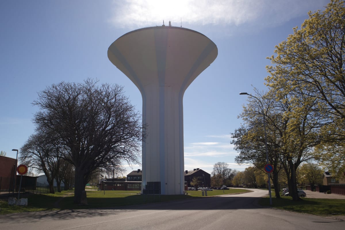 En stor, ljusbrun och vit vattenreservoar, formad som en svamp, står centralt mot en blå himmel med tunna vita moln. Runt omkring syns träd, några med löv och andra kala, samt byggnader och en asfalterad väg framför.