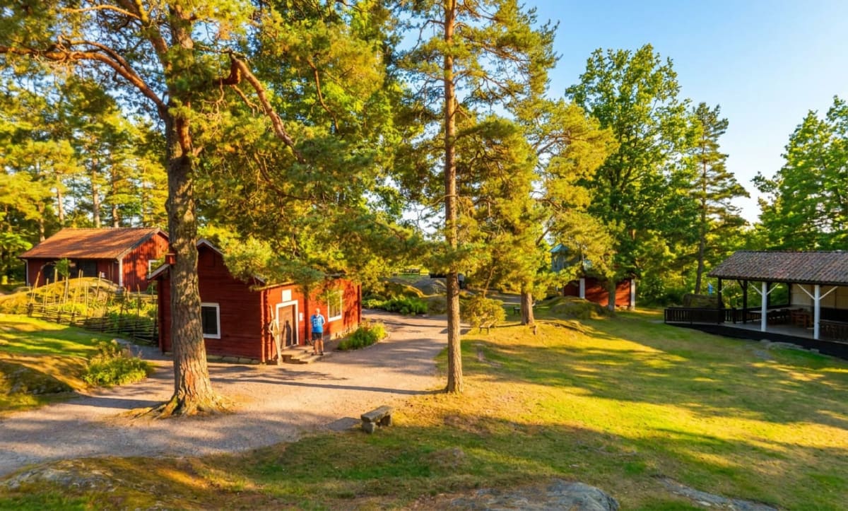 En utomhusbild som visar röda trähus med röda taktegel. På vänster sida finns ett staket av träpinnar och i förgrunden en grusgång. Gröna träd och gräs syns överallt under en blå himmel. Ljuset på sken på byggnaderna och skapar skuggor, vilket indike
