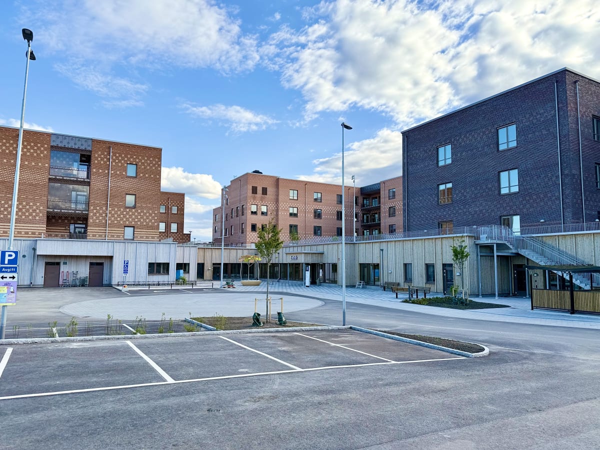 En bild som visar en modern byggnadskomplex med tegel- och träfasader under en blå himmel med vita moln. Framför byggnaderna finns en asfalterad parkeringsplats med vita markeringar och en gångväg. Mellan byggnaderna syns planteringar och en entré so