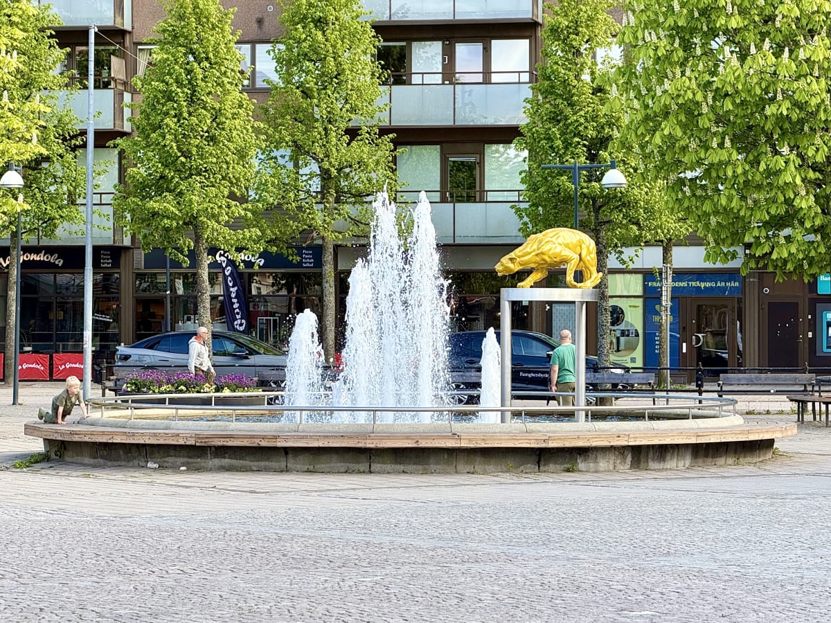 En bild av en stadsmiljö med en stor fontän och en guldfärgad björnstaty i förgrunden. I bakgrunden syns byggnader med reflekterande fönster, träd med ljusgröna löv och några personer. Trädens grenar skymmer delvis byggnaderna bakom.