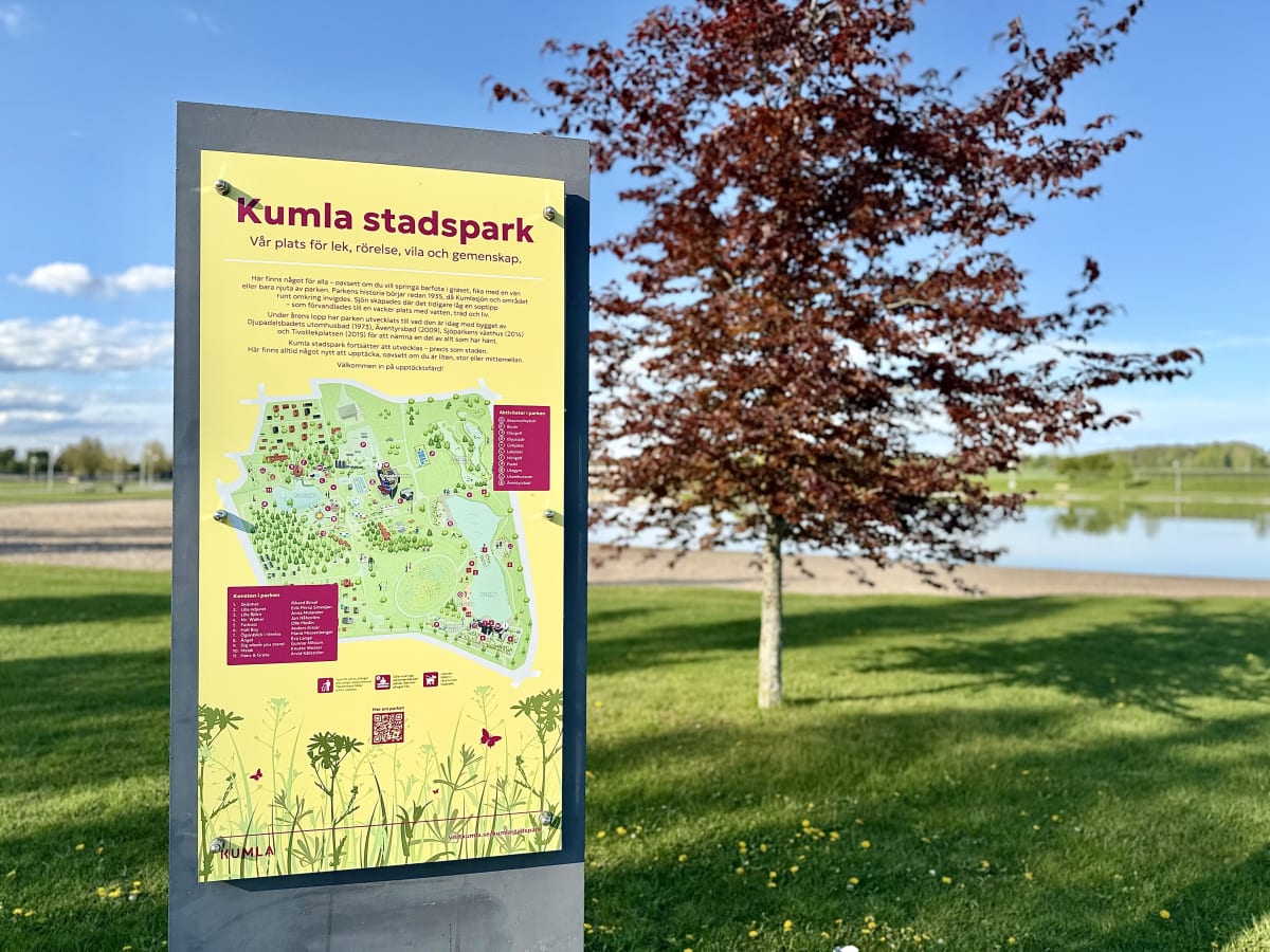 En gul informationsskylt med en karta över Kumla stadspark och text syns på vänster sida. Till höger om skylten finns grön gräsmatta, en lövfällande buske med rödbruna blad, samt en vattenyta och en sandstrand i bakgrunden. En klarblå himmel med få m