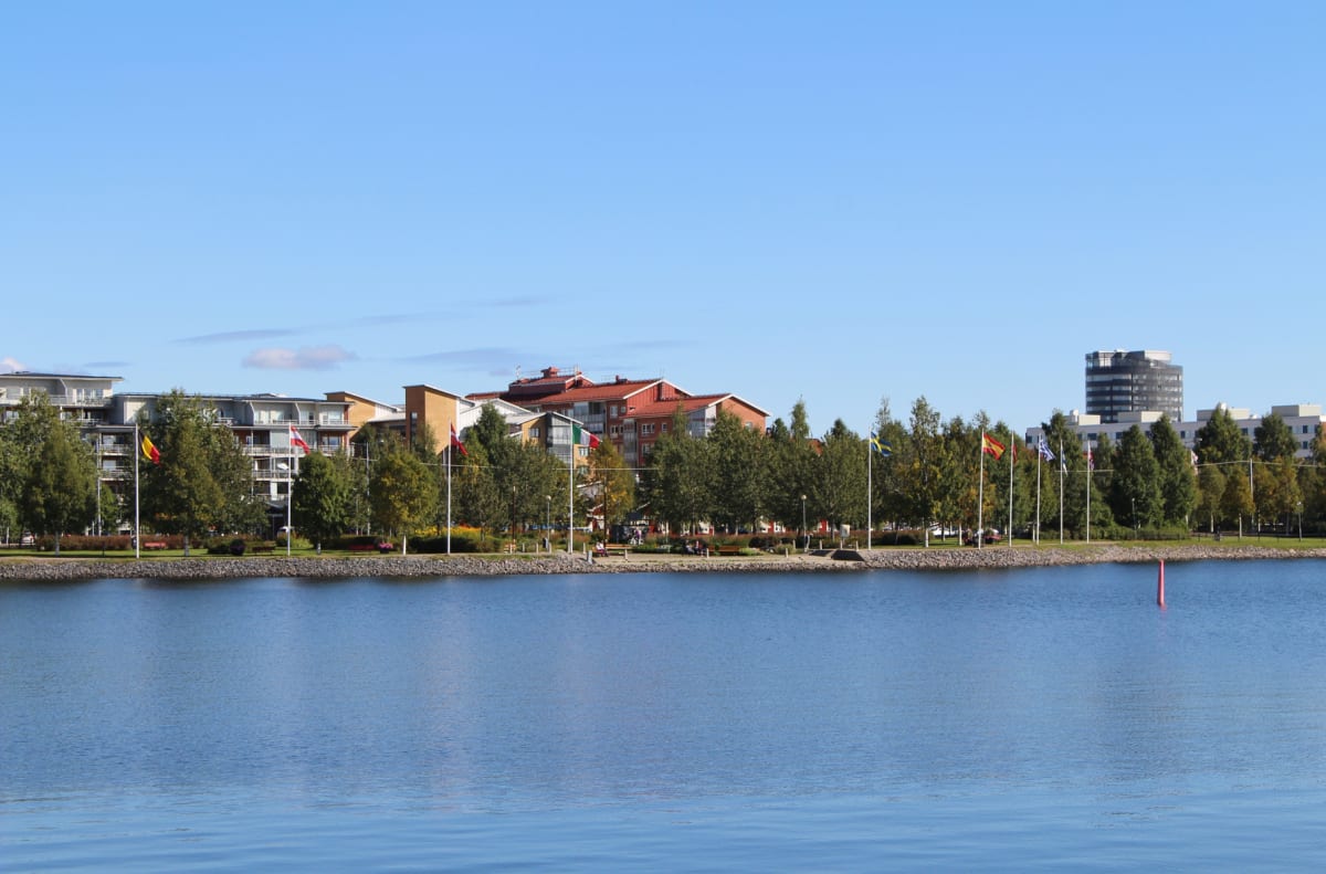 En vidvinkelbild tagen utomhus visar en sjö eller större vattensamling med en horisontlinje av byggnader, träd med gröna löv och flaggstänger med olika nationers flaggor. Den blå himlen är synlig och på vattnet syns en röd boj.
